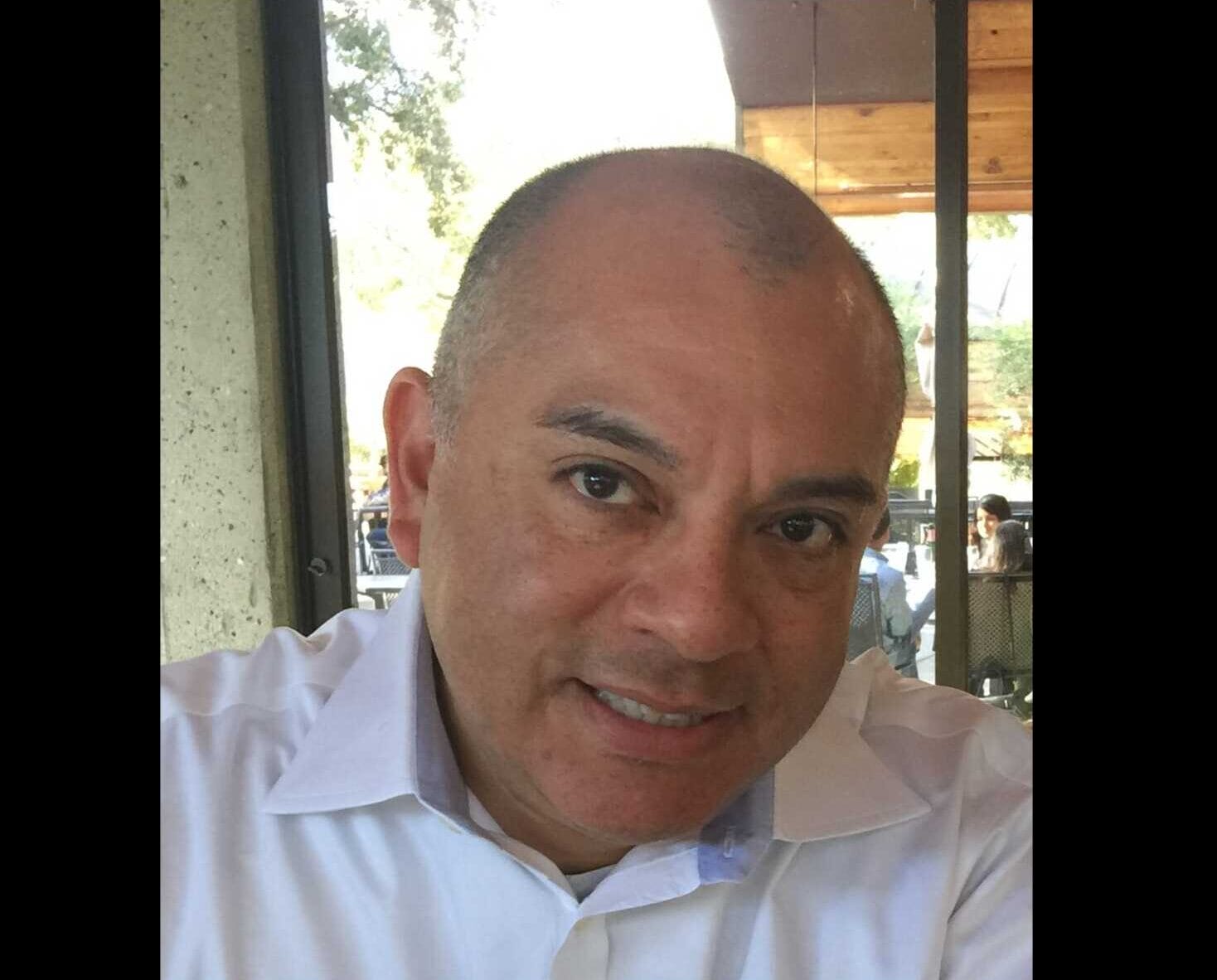 Herndon Town Councilmember Cesar del Aguila (courtesy Cesar del Aguila) | FFXnow Close-up portrait of a smiling man in a white collared shirt, indoors near a window with outdoor cafe seating visible behind him, looking at the camera.