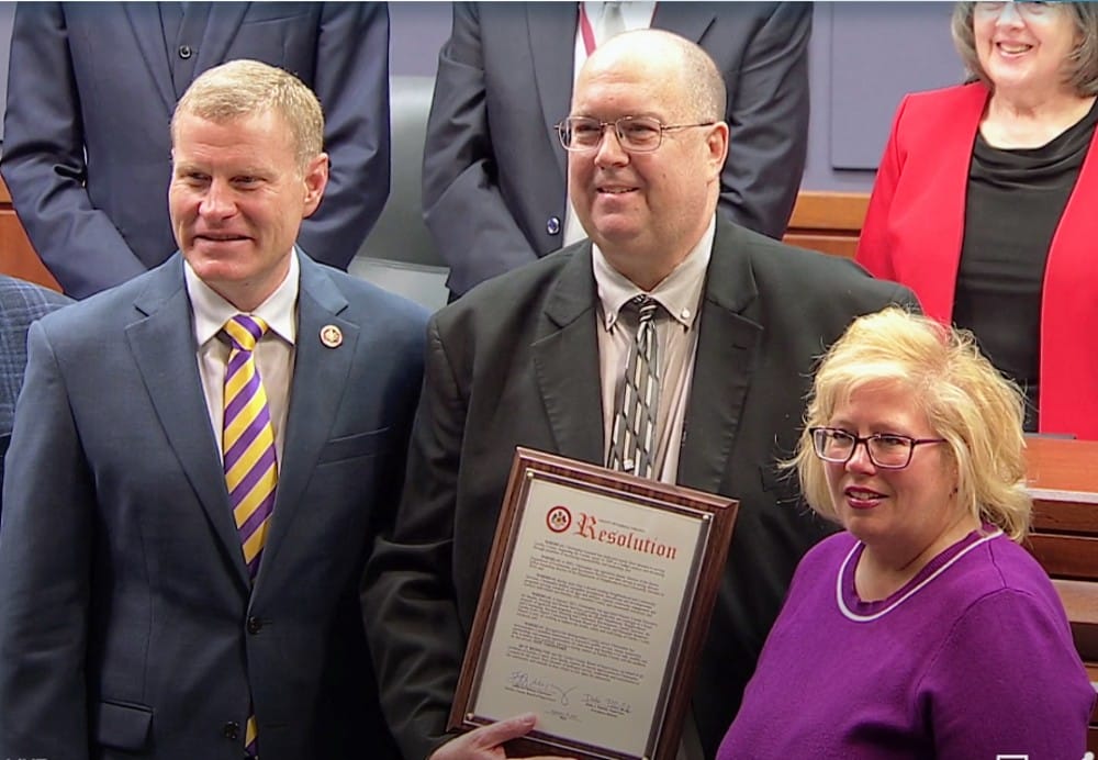 Christopher Leonard during presentation at Board of Supervisors' meeting (screenshot via Fairfax County) | FFXnow Three adults in business attire pose for a photo; the center man holds a framed document, flanked by a man in a blue suit and a woman in a purple sweater.