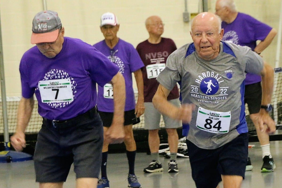 Competitors in the Northern Virginia Senior Olympics 2026 men's 60-meter running event (staff photo by Scott McCaffrey) | FFXnow Two elderly men lead an indoor race, one in a purple shirt with bib 7 and the other in a gray shirt with bib 84, leaning forward in sprinting motion.