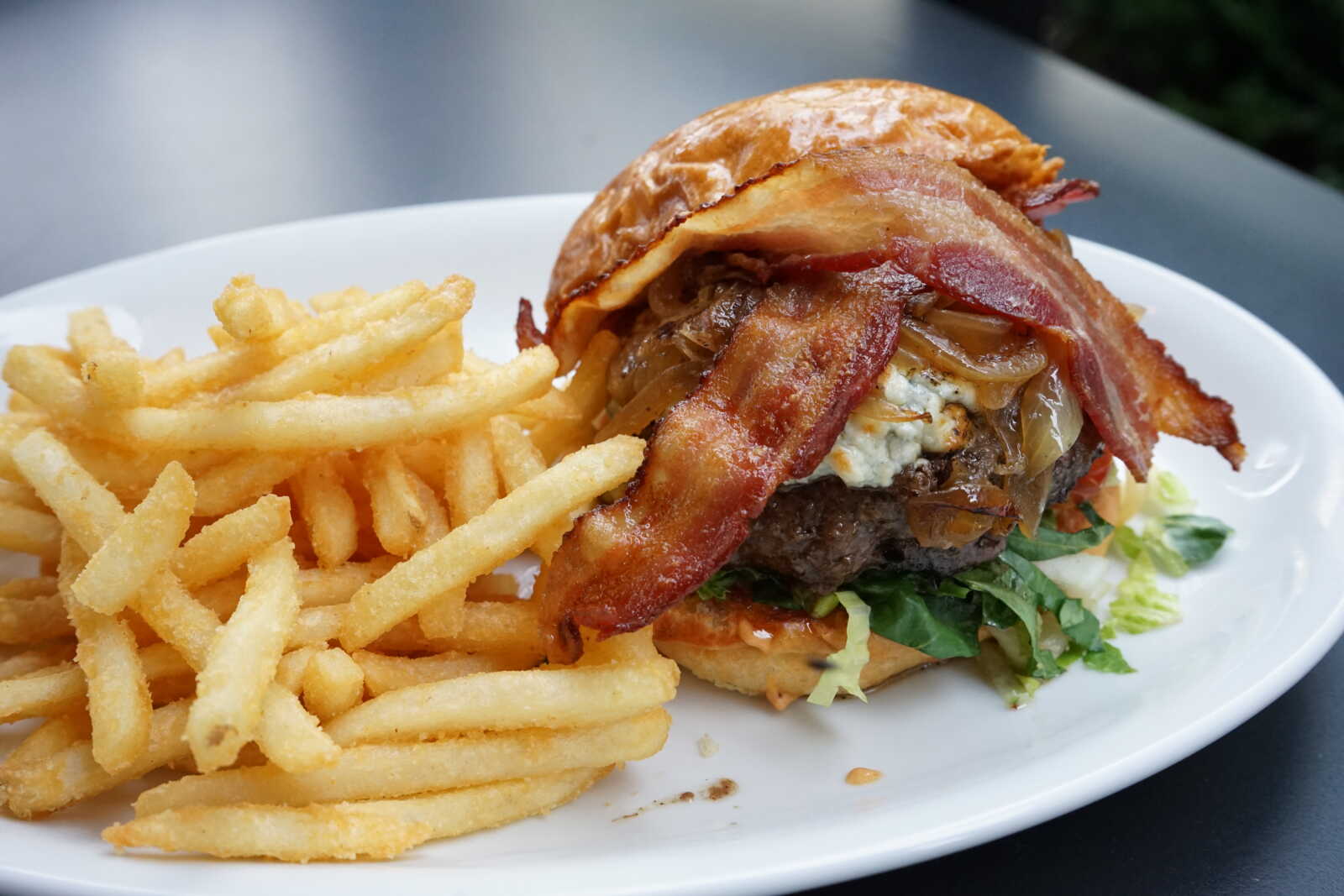 Burger with bacon, onions, and greens on a toasted bun, served with fries on a white plate.