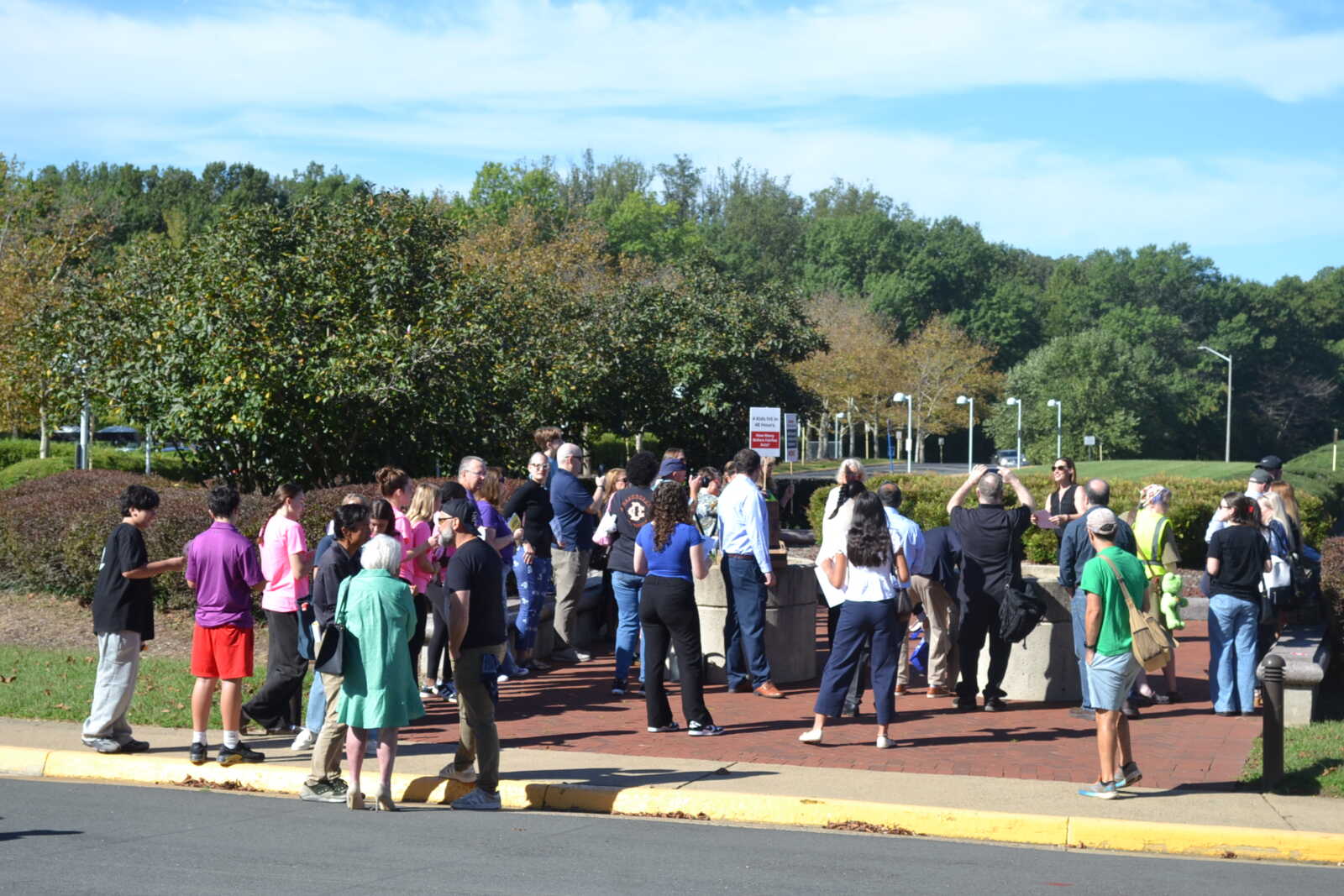 A crowd of people gathered on a brick plaza beside hedges and trees on a sunny day in a park setting fin outdoor gathering.