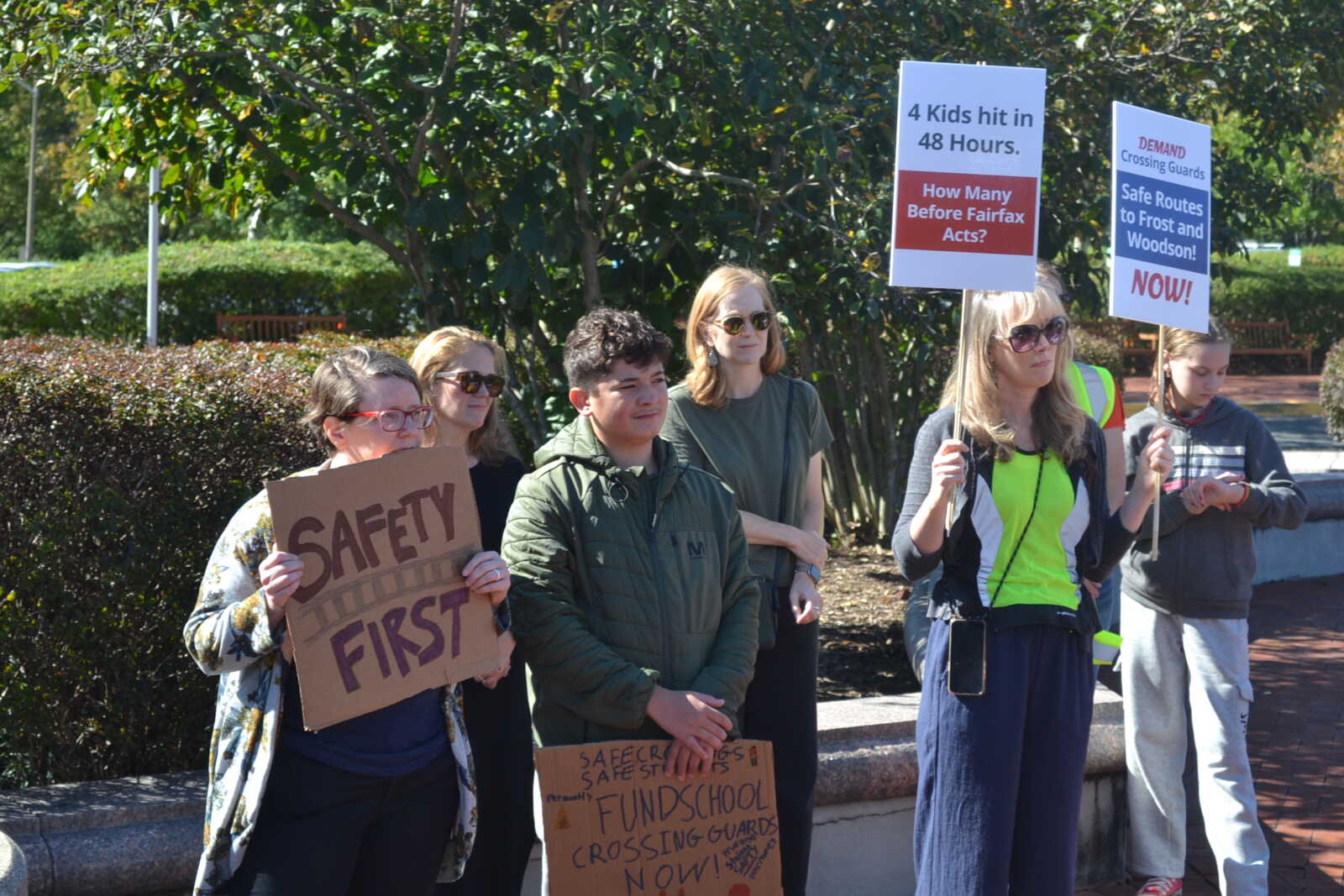 Group of protesters outdoors holding signs about school safety, including a large 'SAFETY FIRST' cardboard sign and two vertical posters demanding safer routes.