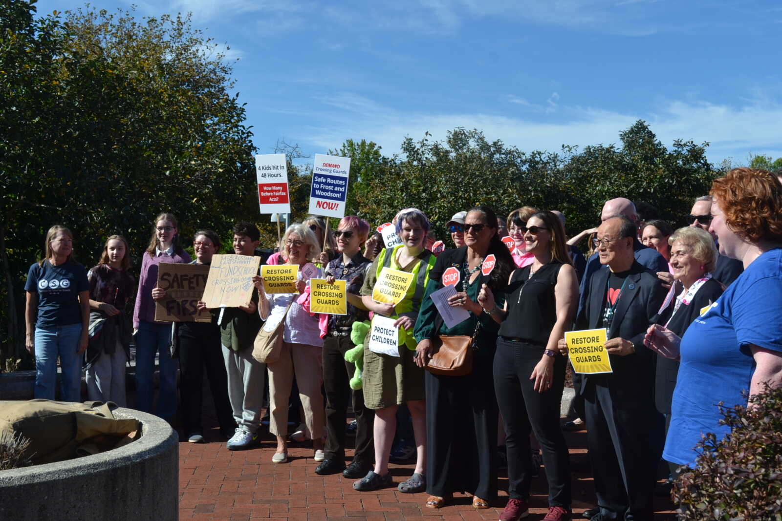 Group of people at a peaceful protest holding signs about school crossing guards outdoors on a sunny day.