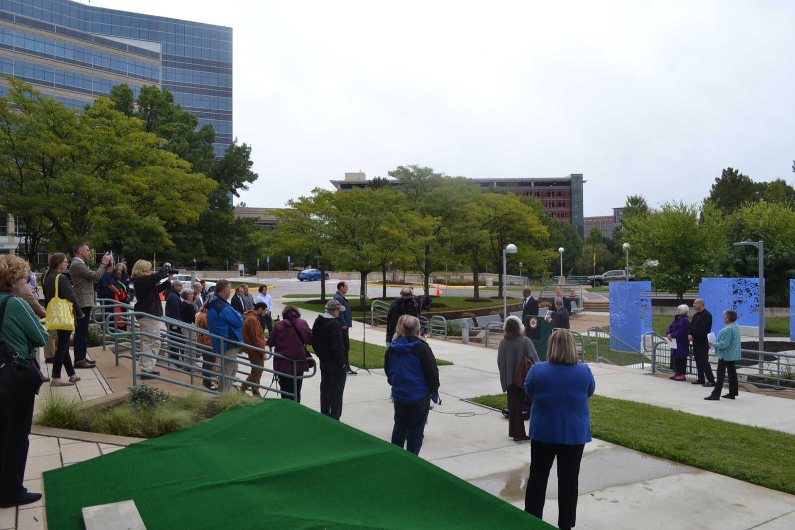 Crowd of people gathered outdoors listening to a speaker at a ceremony, with trees, modern buildings, and blue display panels in the background.