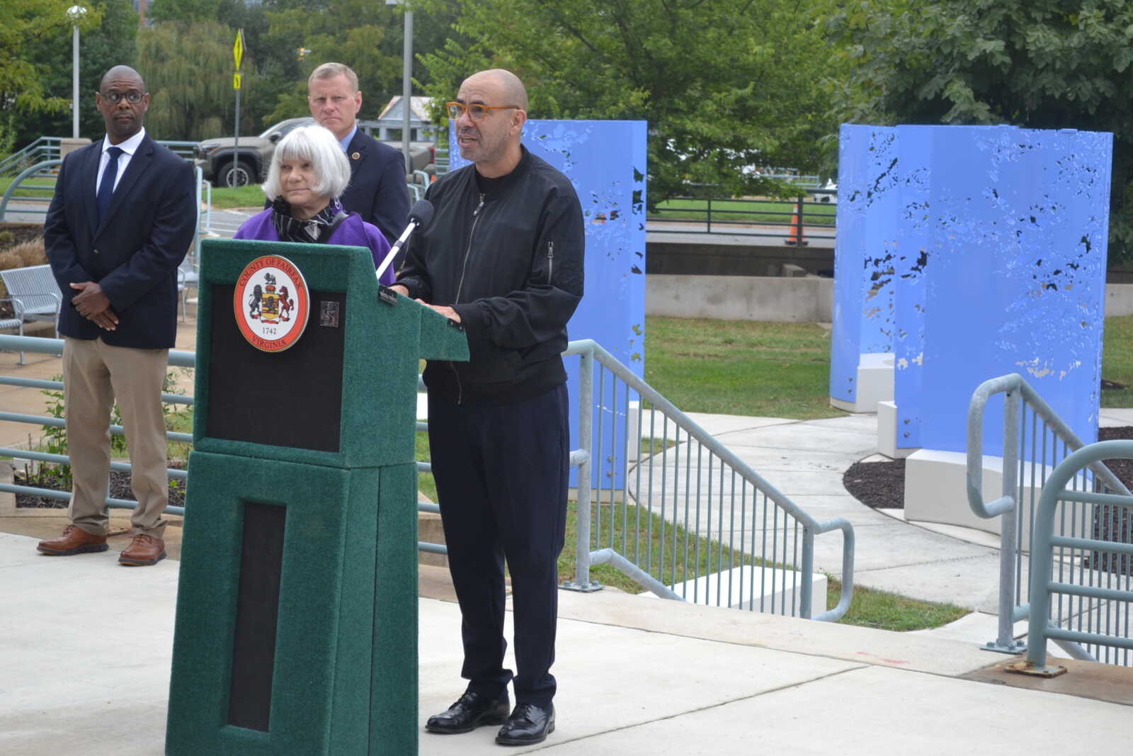 Man in a black jacket speaks at a green podium with a city seal, flanked by two adults in business attire outdoors behind him.