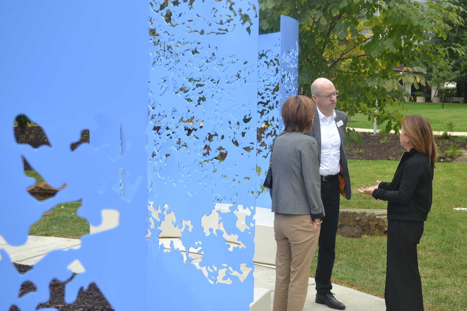 Three professionals in business attire converse beside a blue perforated sculpture in a park.