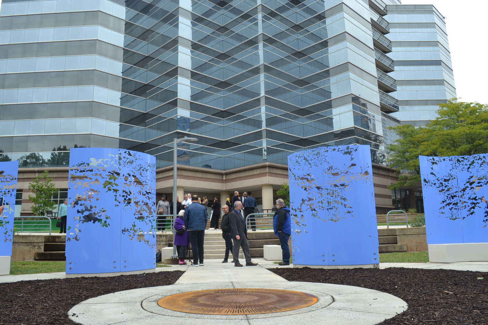 Group of people gathered outside a modern glass office building, standing near blue perforated panels in a plaza courtyard.