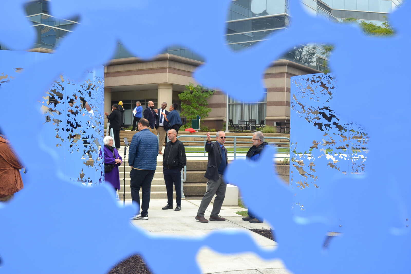 Group of people chatting and mingling on a plaza in front of a modern office building, seen through blue decorative cutout panels in the foreground.