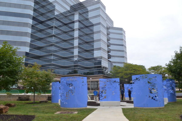 Public park with a glossy glass office building in the background and blue perforated cylinder sculptures in the foreground.
