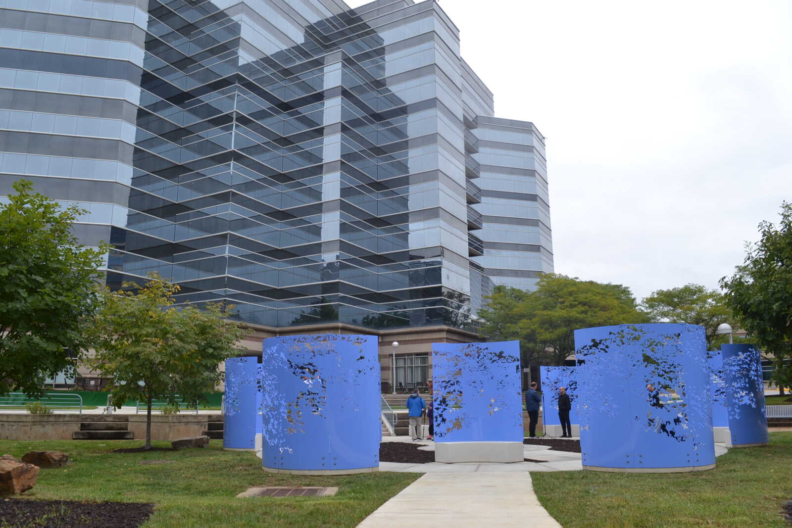 Public park with a glossy glass office building in the background and blue perforated cylinder sculptures in the foreground.