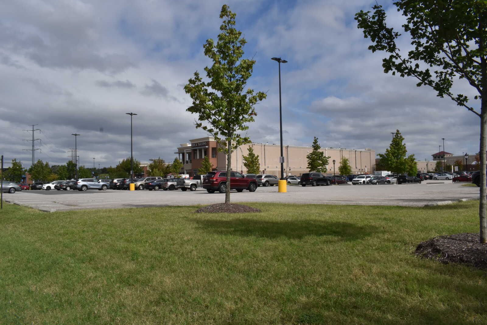 Parking lot in front of a beige shopping center with many parked cars and a grassy foreground under a cloudy sky