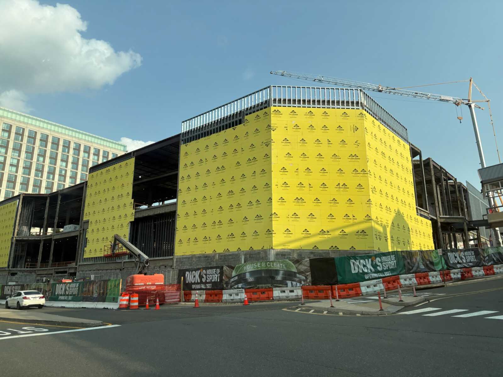 Dick's House of Sport under construction at Tysons Corner Center (photo by Amy Woolsey) | FFXnow Under-construction building wrapped in yellow weather barrier with a crane overhead and city street in front.