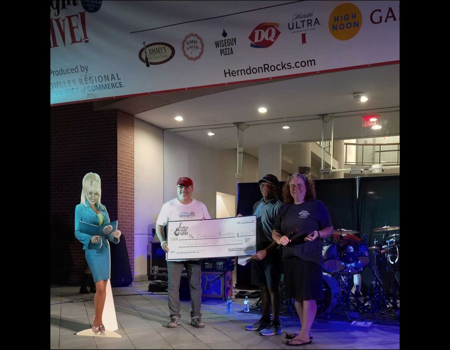 Friday Night Live! founder Doug Downer and chair Laura B. Poindexter present a check for 0 to Kurt Rose support Dolly Parton's Imagination Library initiative in Herndon (photo by Any Odhner) | FFXnow Four people on a stage holding a large ceremonial check at a community event, with a life-size cutout and a drum set in the background.