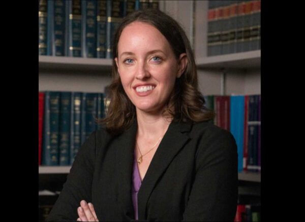 Professional woman in a black blazer, smiling, with bookshelves in the background.