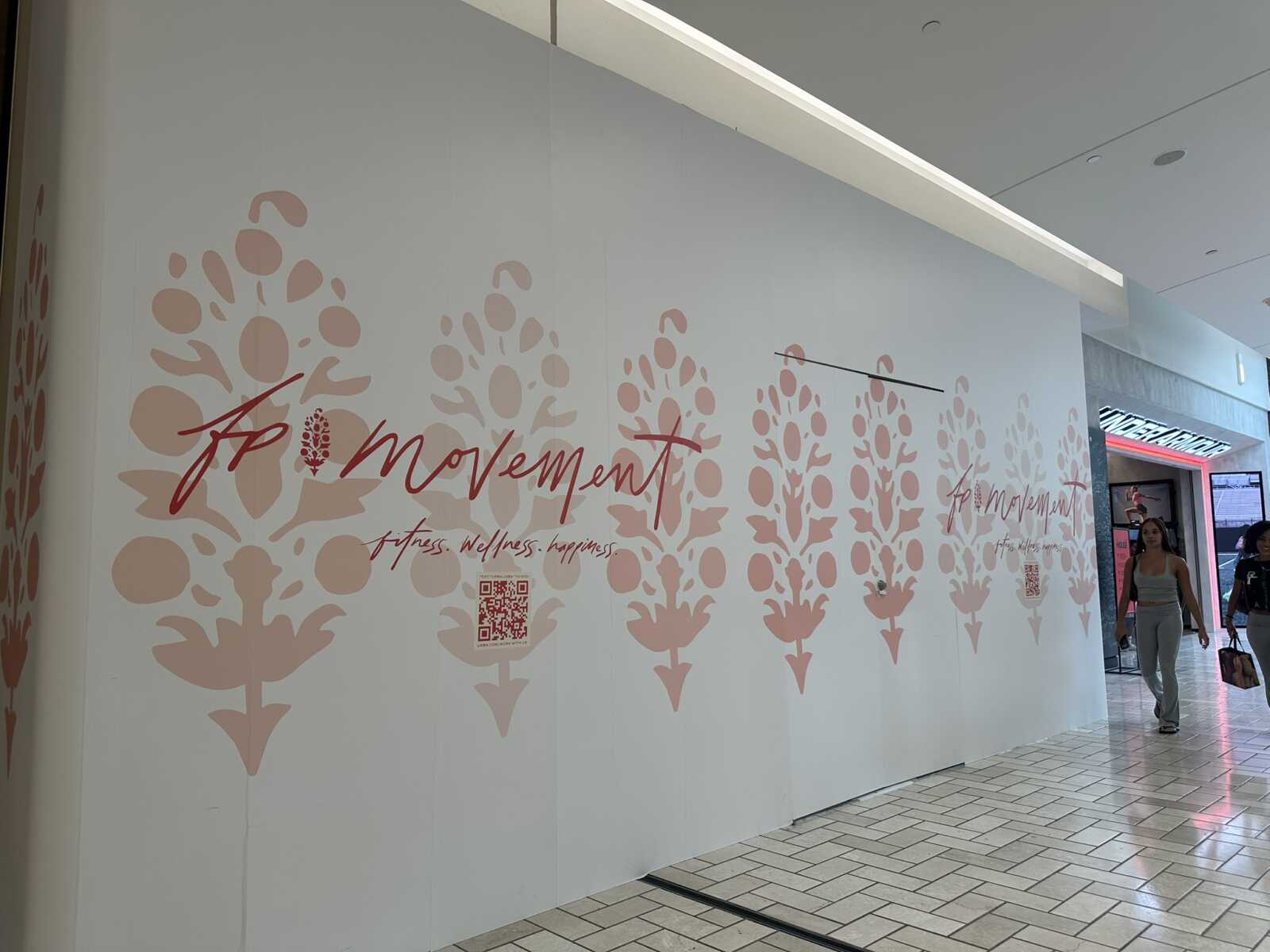 White interior wall with large pink floral motifs and 'Momentum' fitness text; two shoppers walk by in a mall corridor.