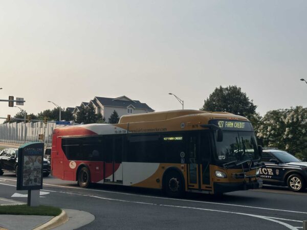 Yellow and red Fairfax Connector bus stopped at an intersection with a police car beside it; destination sign reads '427 Farm Credit' on the front. A Capital One Center bus stop sign is in the foreground.
