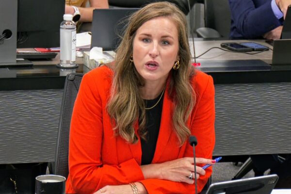 Woman in a red blazer speaks into a microphone at a conference table a panel setting.