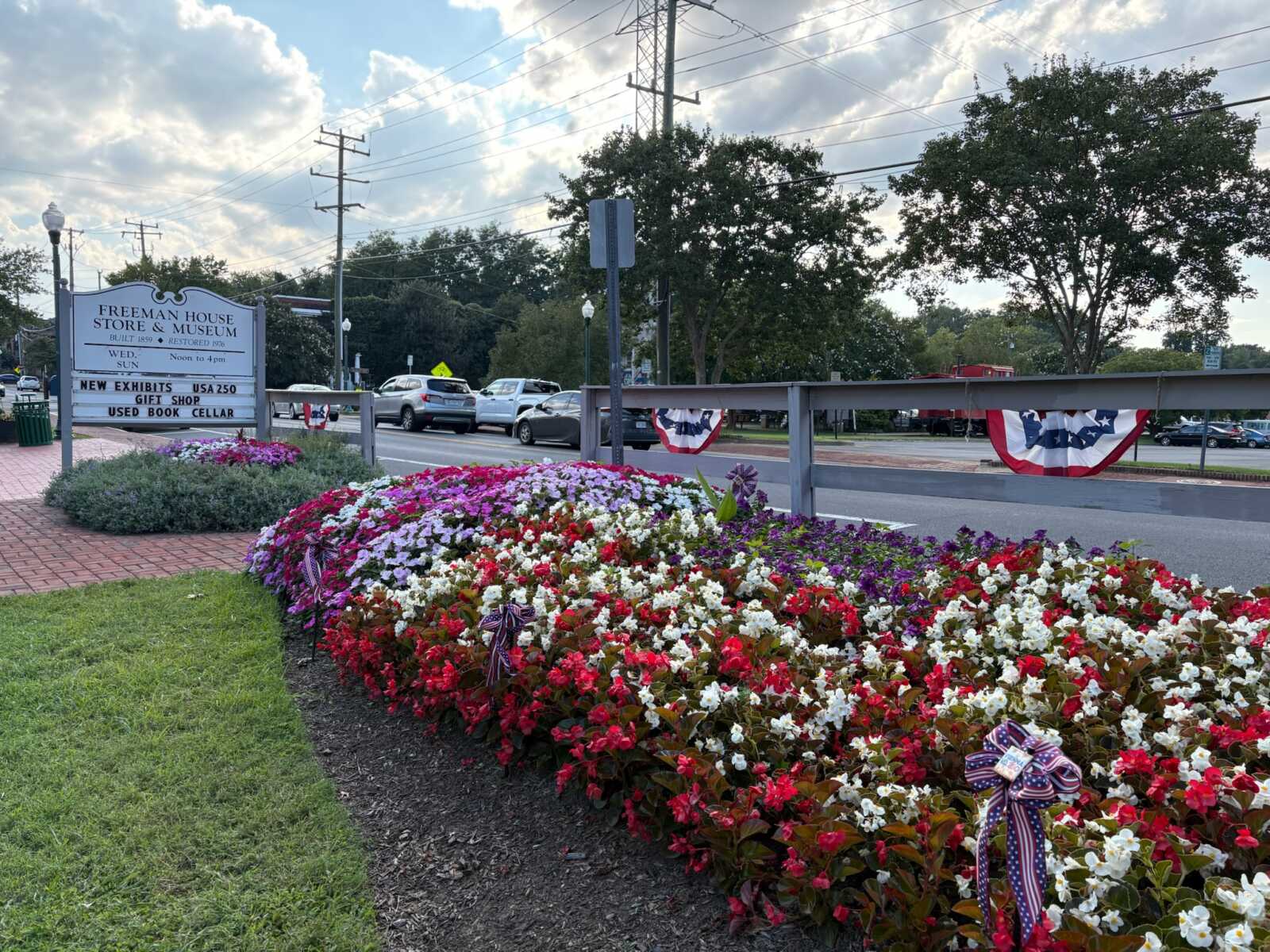 Flowers with patriotic bunting outside the Freeman Store and Museum in Vienna (photo by Amy Woolsey) | FFXnow Flower bed of red, white, and purple blooms beside a sidewalk and a Freeman House Store & Museum sign with patriotic bunting along the railing in the street scene.