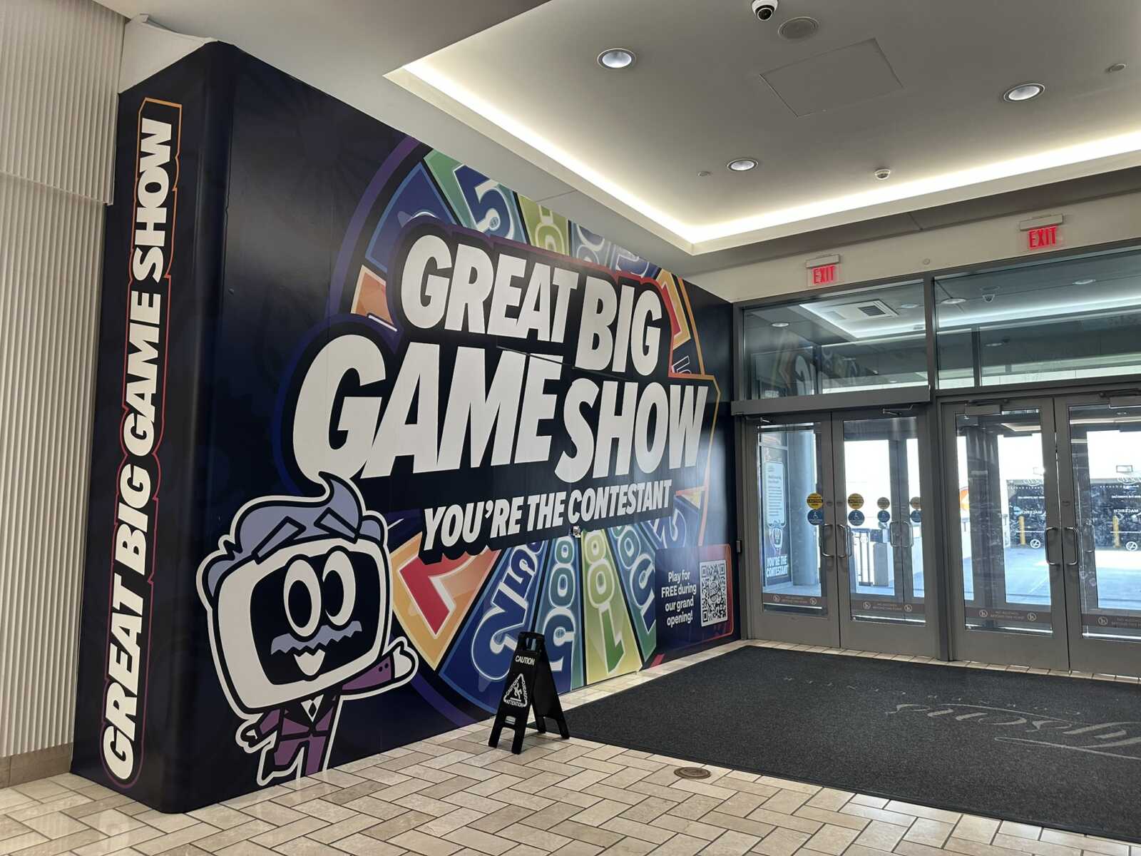 Colorful wall mural says 'Great Big Game Show' with a cartoon mascot, at a mall entrance near glass doors.