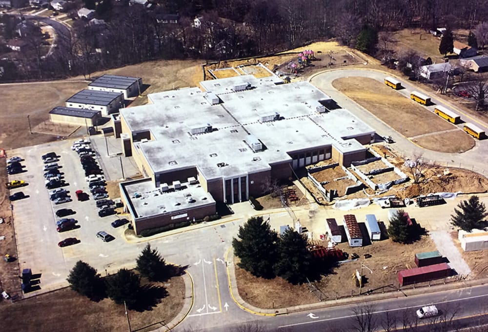 Aerial view of a large flat-roofed building with ongoing construction on the side, surrounded by a parking lot and yellow school buses nearby.