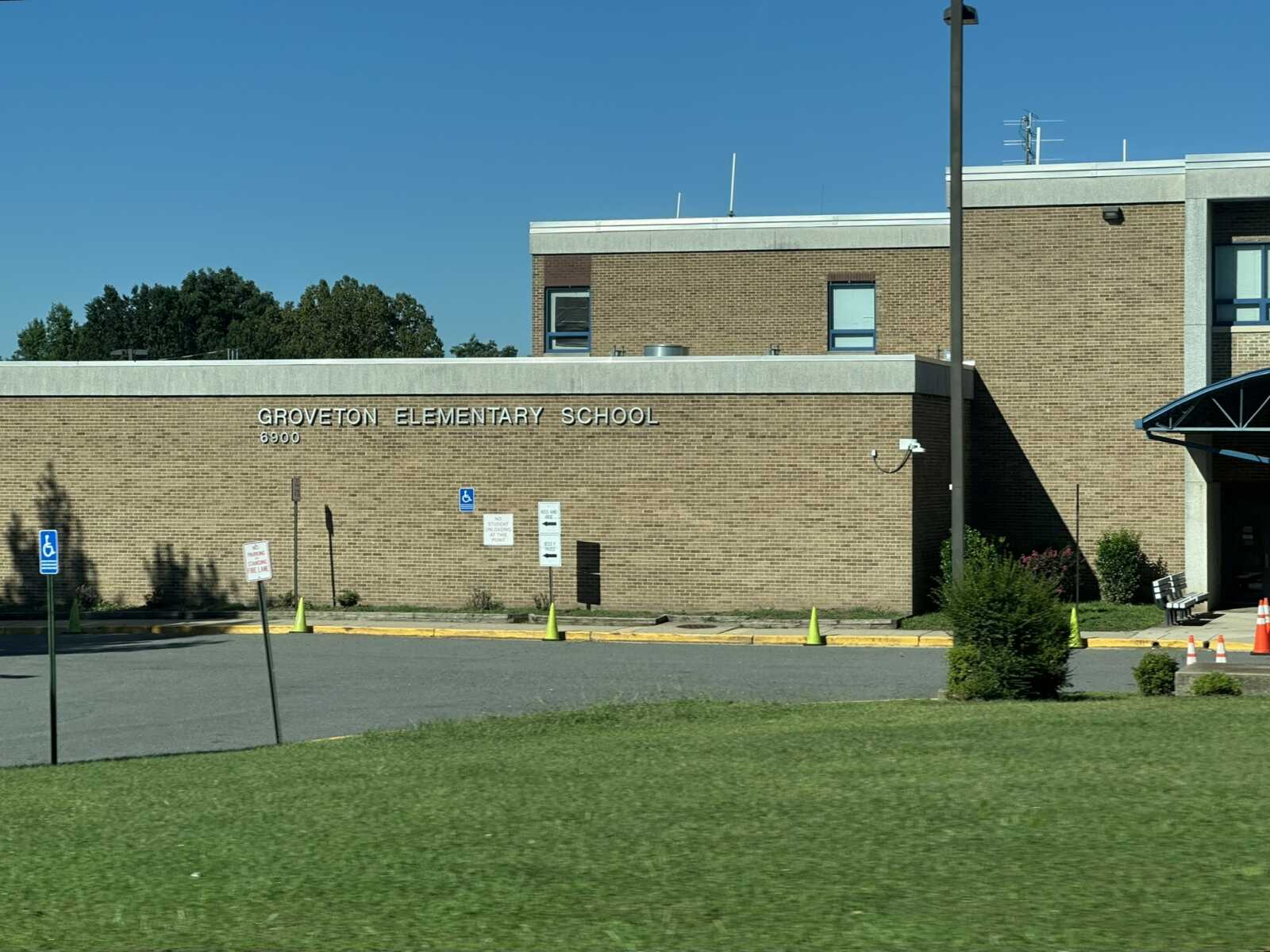 Front view of Groveton Elementary School—brick building with the school name on the facade, a clear blue sky, and a parking lot with handicap signs and cones in front.