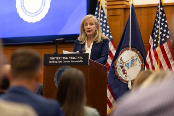 A woman with blonde hair, wearing a suit, stands at a podium surrounded by people. The Virginia state flag is visible on a screen in the left corner behind her.