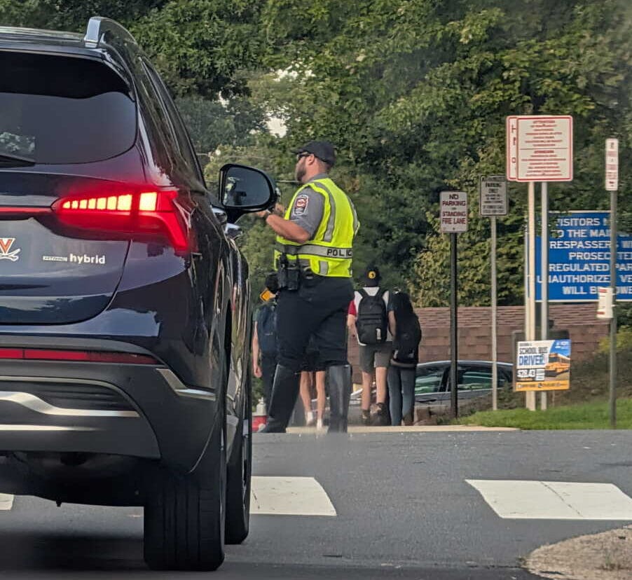 Police officer in a reflective vest talks to pedestrians at a crosswalk beside a dark SUV.