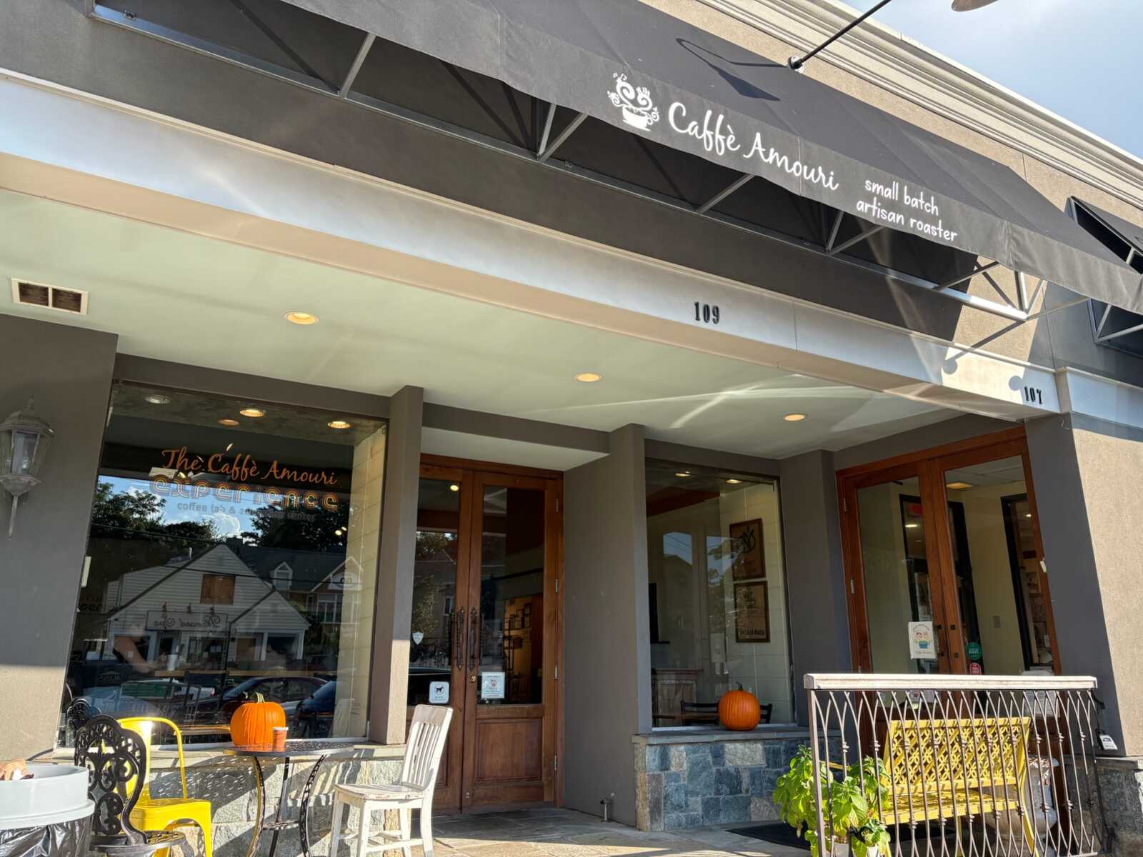 Caffe Amouri storefront in Vienna (photo by Amy Woolsey) | FFXnow Exterior view of The Caffé Amouri coffee shop with a black awning, glass windows, and wooden doors; pumpkins decorate the outdoor seating area on a sunny day.