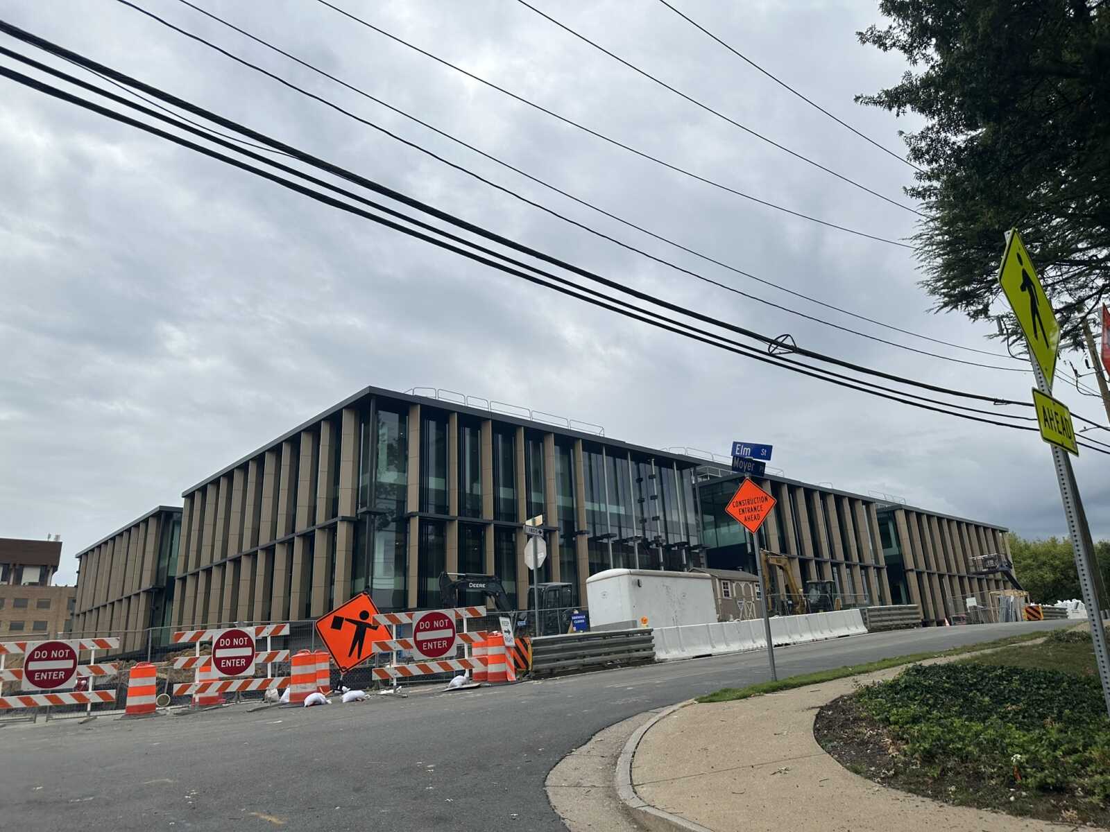 Construction site blocks a street in front of a modern glass-and-metal building; orange barriers and Do Not Enter signs are visible with traffic signs nearby.