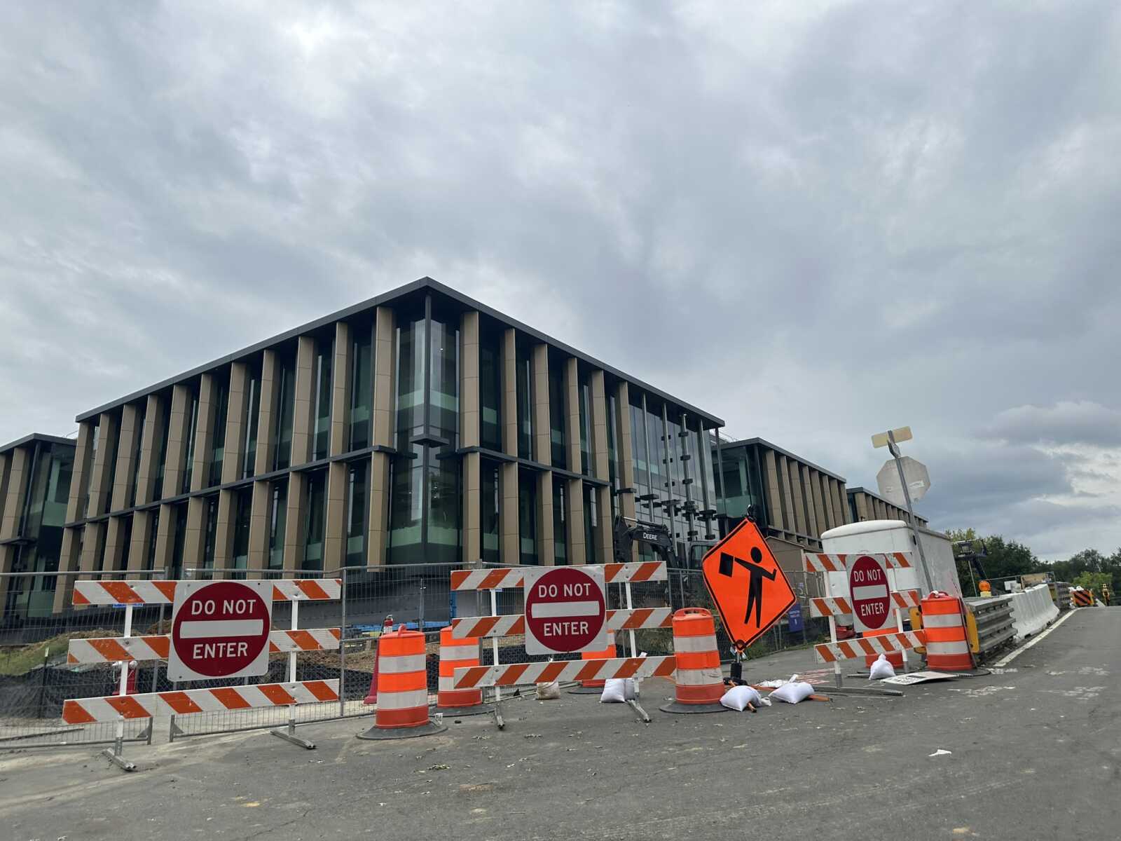 Construction barriers and orange cones block a street in front of a modern glass-and-wood building under a cloudy sky, with 'Do Not Enter' signs warning of a restricted area.