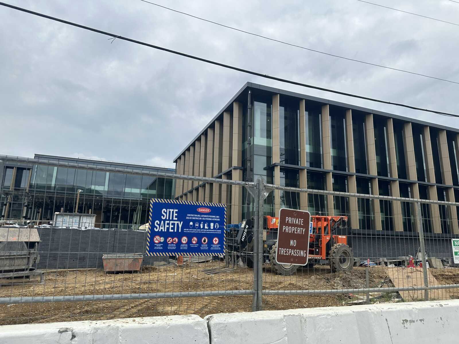 Construction site with a modern glass-and-beam building behind a chain-link fence and safety signs.