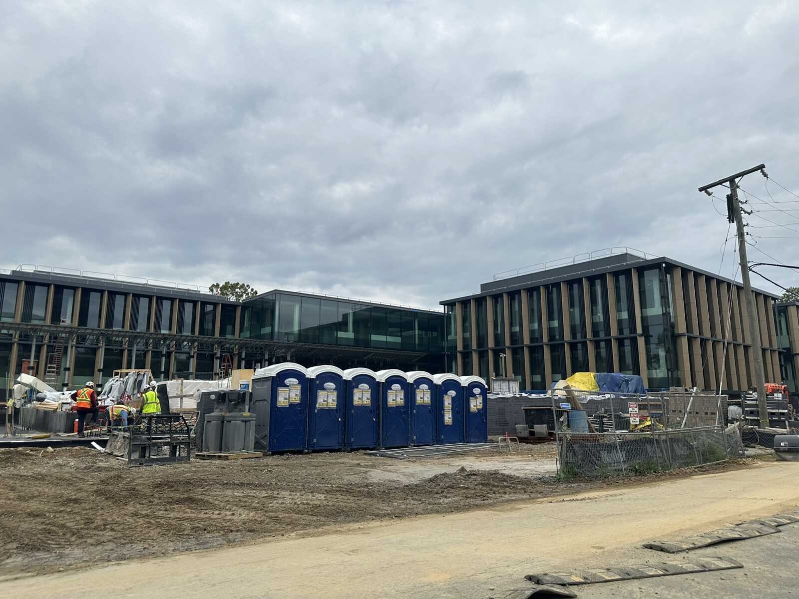 Construction site in front of a modern glass-and-beam building; portable toilets and workers in high-visibility vests.