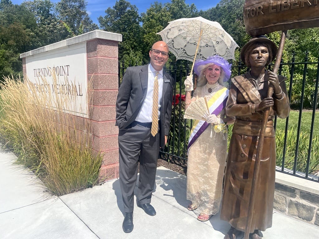 Man in suit posing with a woman in a vintage lace dress under a lace parasol, beside a bronze statue at a memorial entrance.