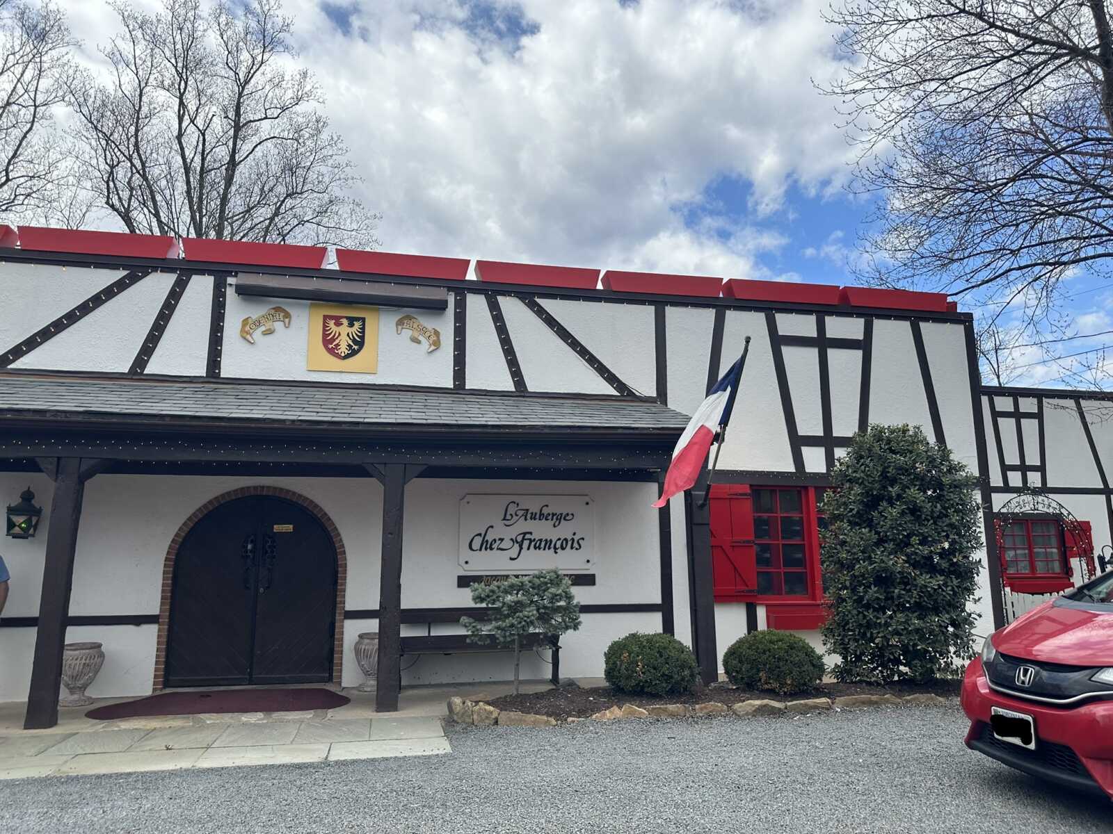 Exterior of a Tudor-style restaurant with black timber framing, white walls, and red shutters, plus a French flag outside.