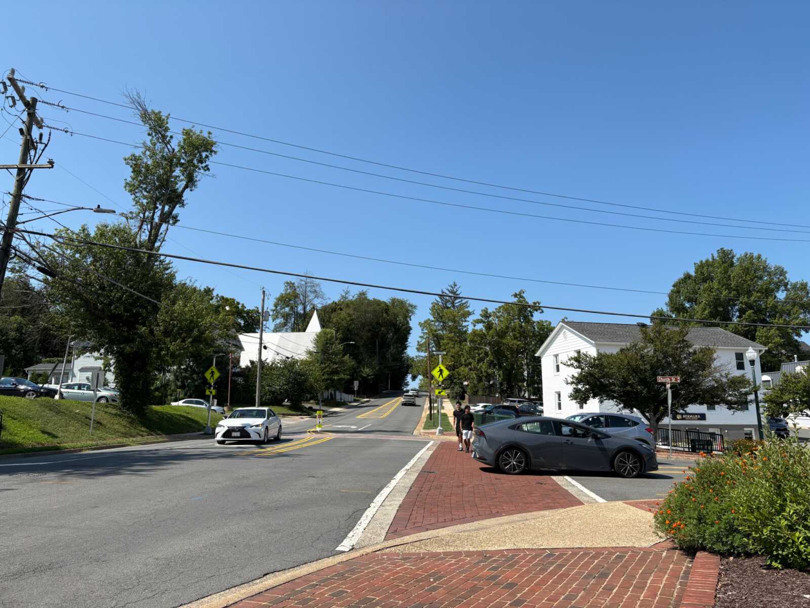 Sunny suburban intersection with brick sidewalk, crosswalk signs, and parked cars along a tree-lined street.