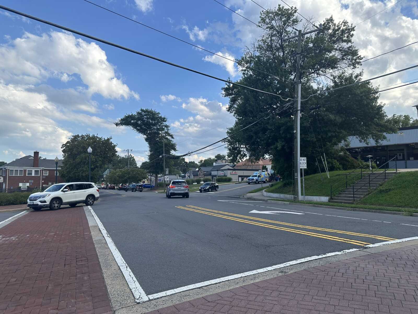 Two-lane street at a small-town intersection with brick sidewalks, parked white SUV, and leafy trees under a blue sky with clouds.