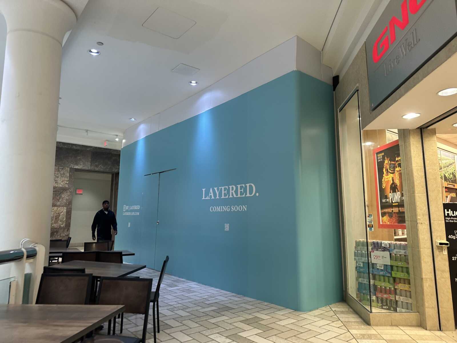 Mall corridor with a teal construction wall reading 'LAYERED. COMING SOON'; foreground has tables and chairs, right side shows a storefront with posters.
