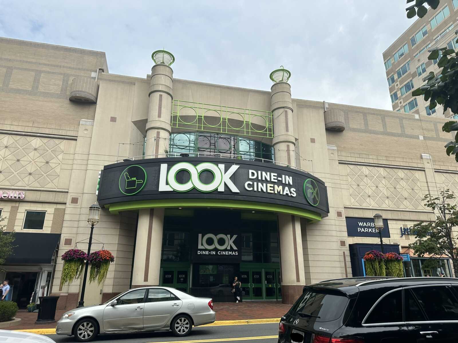 The entrance to Look Dine-In Cinemas at Reston Town Center (staff photo by Angela Woolsey) | FFXnow Exterior of Look Dine-In Cinemas with a curved black marquee and green logo, beige building, and parked cars in front.