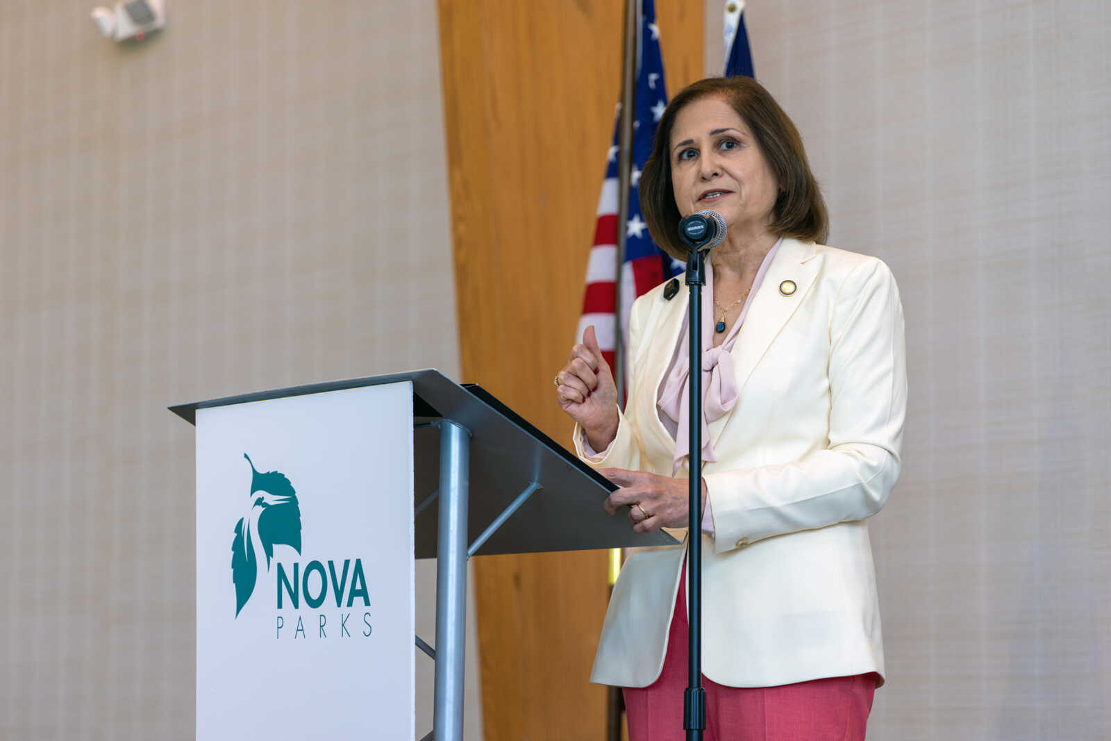 Woman in a light blazer speaking at a podium with NOVA Parks logo, American flag in the background.
