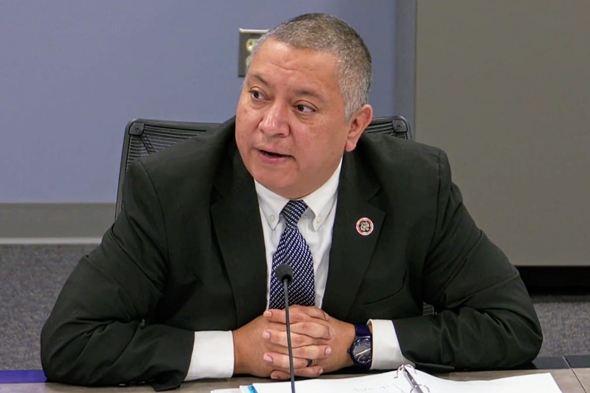 Mason District Supervisor Andres Jimenez (screenshot via Fairfax County) | FFXnow Man in a suit seated at a table with a microphone, speaking during a formal meeting or hearing.