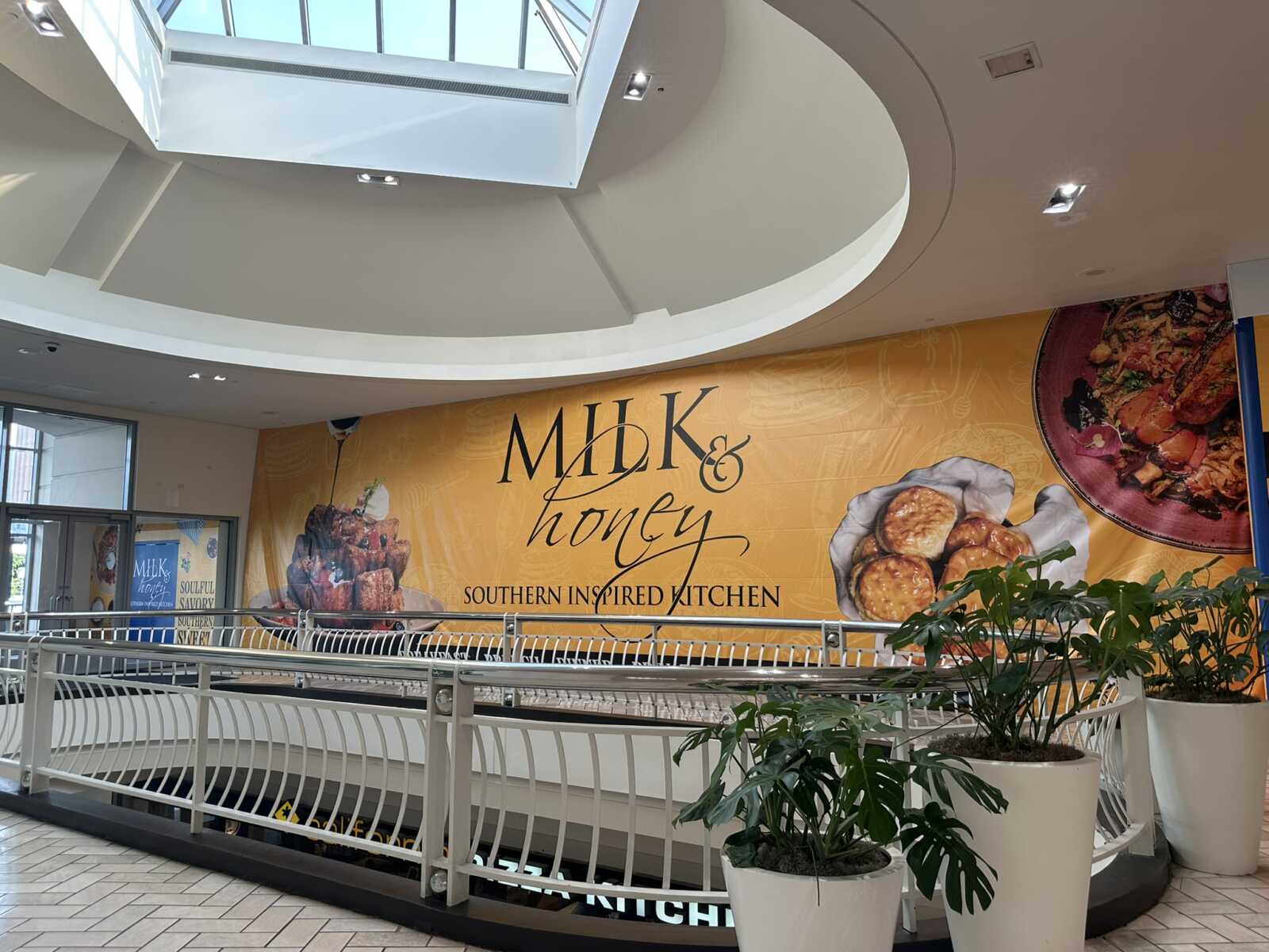 Indoor mall atrium with a large yellow MDK & honey advertisement wall and potted plants in white planters the foreground.