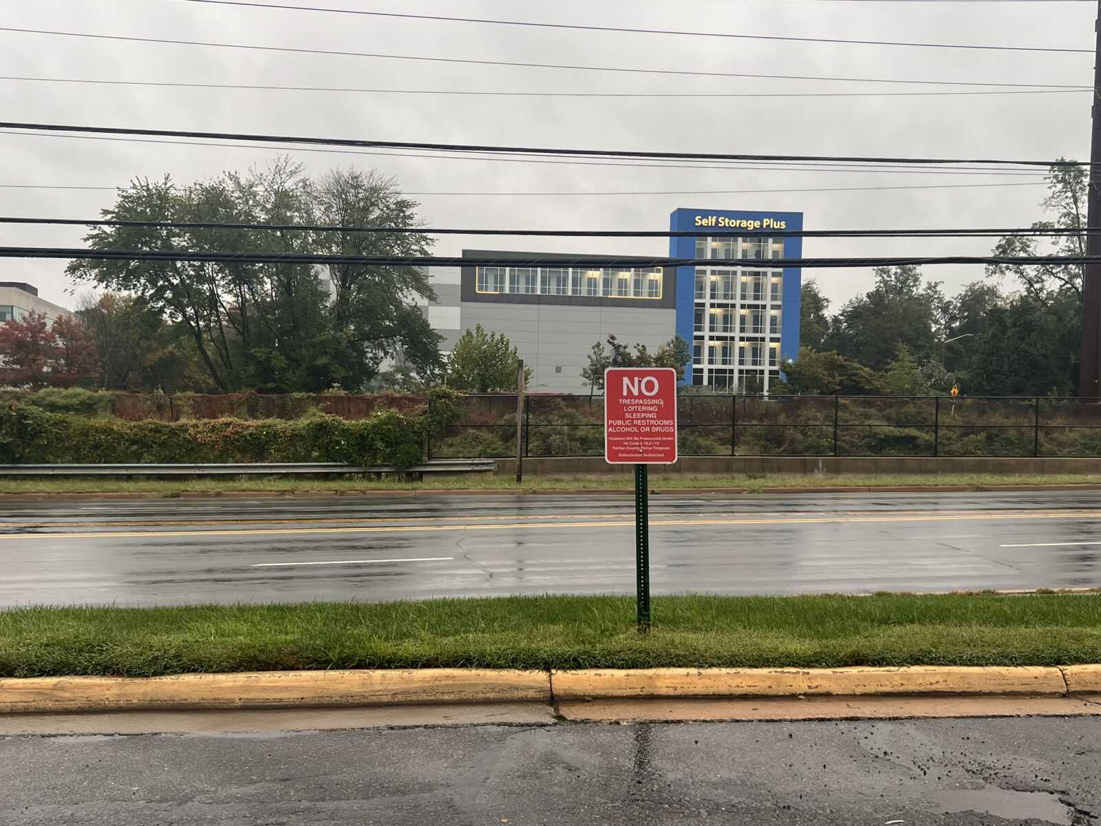 Red 'NO TRESPASSING/Al COHOL OR DRUGS' sign on a post by a wet road, with a chain-link fence and a modern Self Storage Plus building in the background.