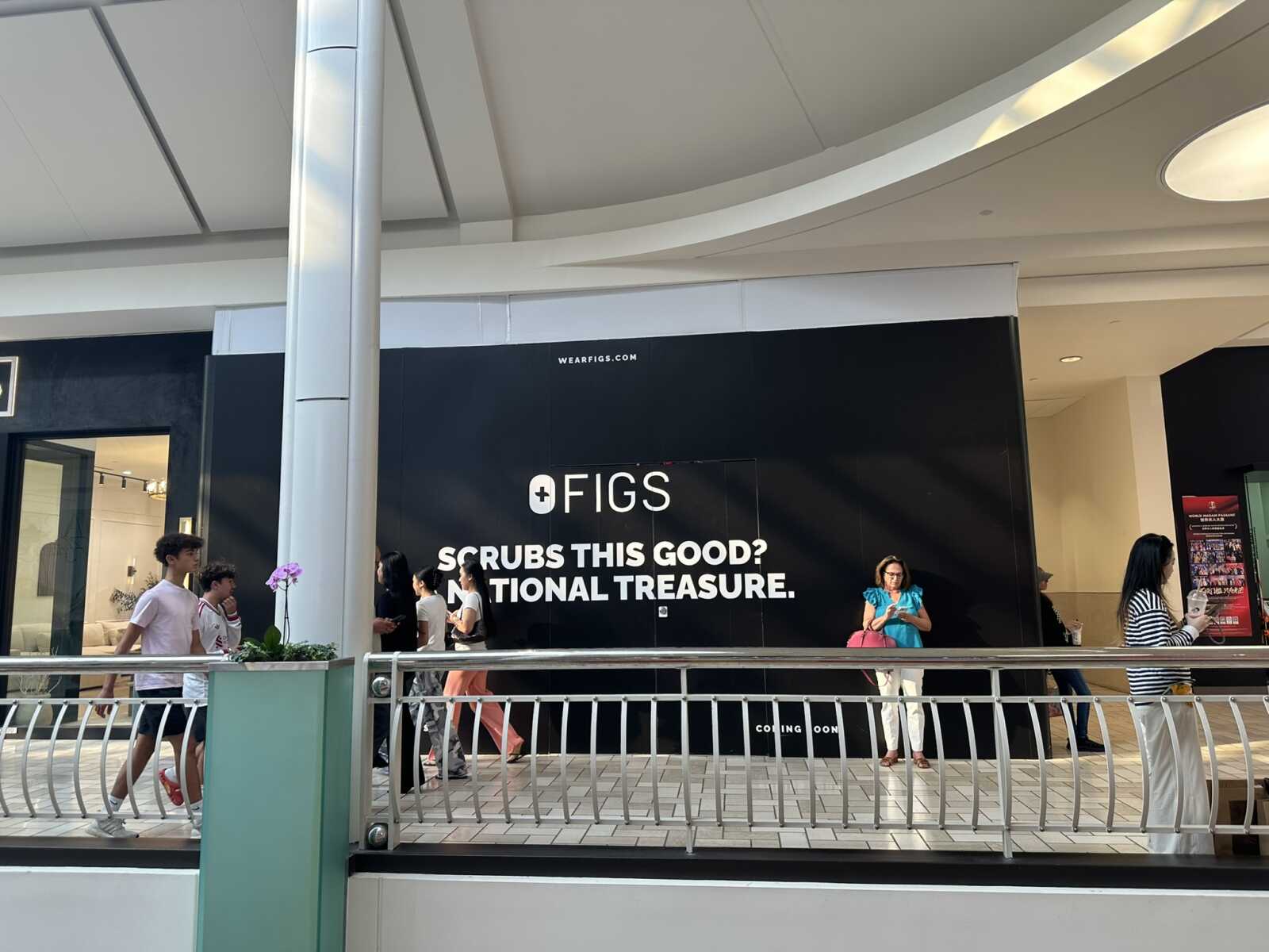 Shoppers pass a large black storefront ad in a mall that reads 'OFIGS SCRUBS THIS GOOD? NATIONAL TREASURE'.