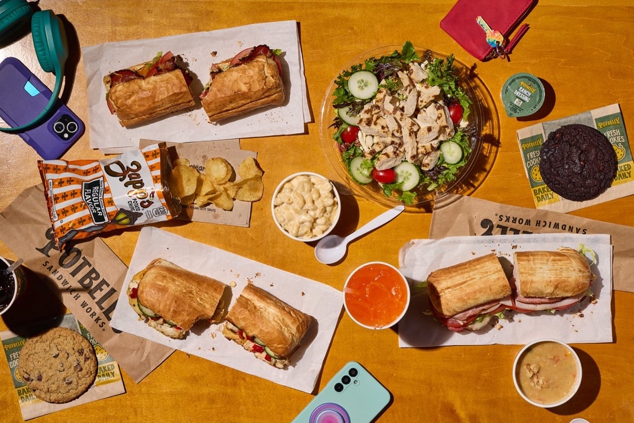 Overhead view of a table full of sandwiches, a large salad, chips, a cookie, and cups of dips and sauces