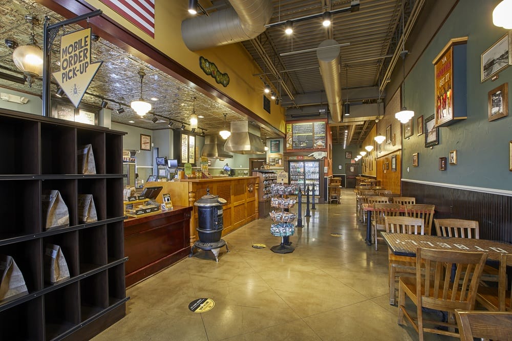 Interior of a cozy cafe with a long wooden counter, tables and chairs along the wall, and warm lighting fixtures.