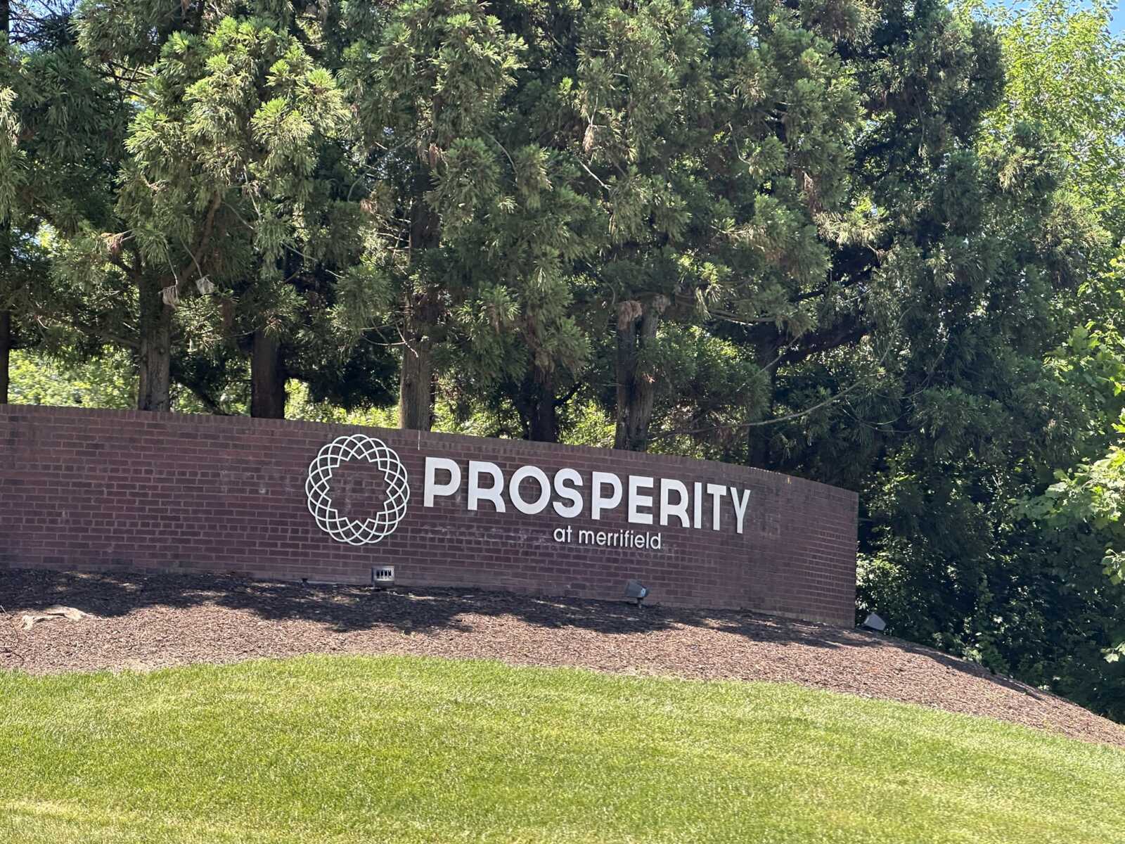 Brick sign reading “PROSPERITY at merrifield” with a white circular geometric logo, set against green trees.