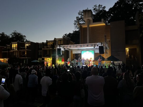Outdoor evening concert with a band playing on a lit stage in front of a brick building and a large attentive crowd.