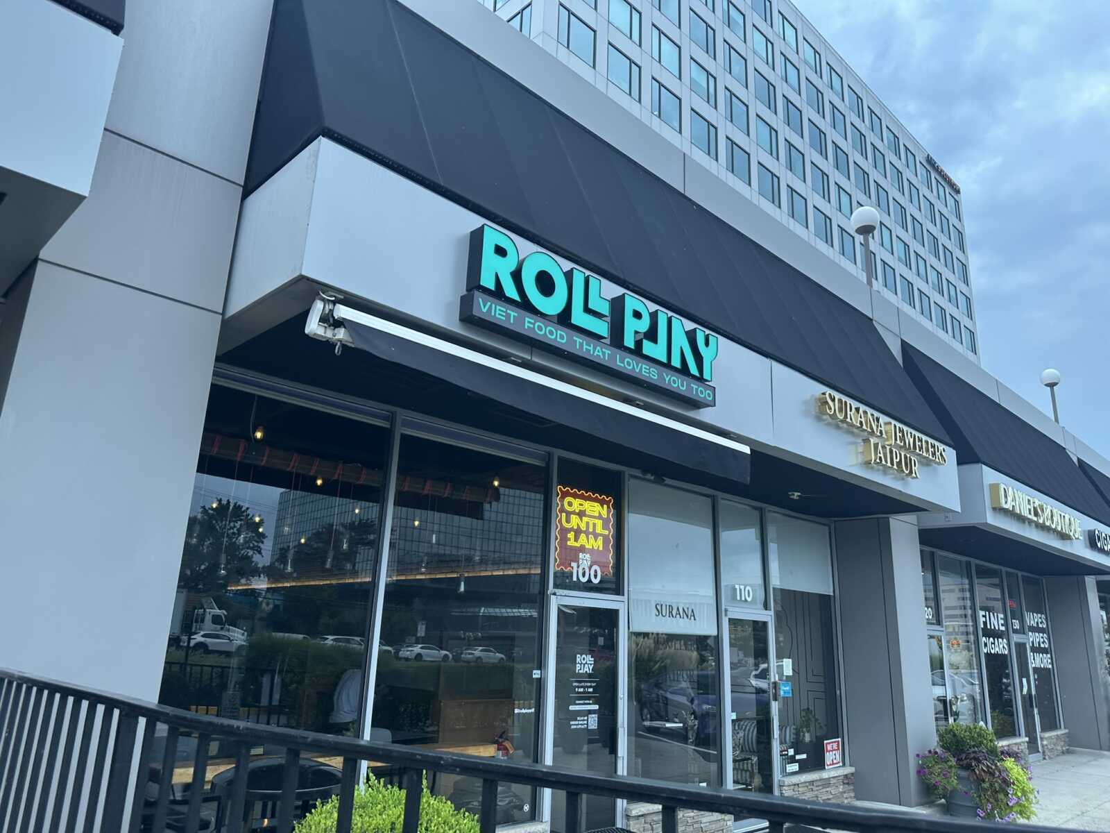 Modern strip mall storefronts with glass windows and a turquoise neon sign reading 'ROL PNY' above the entrance.