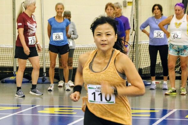 Female runner in an orange tank top and bib 110 sprints on an indoor track while a group of onlookers with race bibs watches in the background.