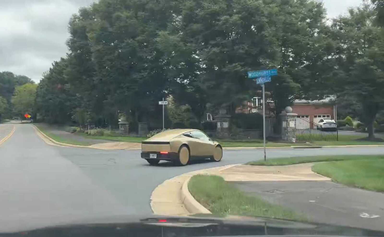 Beige, bubble-shaped concept car turning at a suburban intersection with trees and houses in the background.